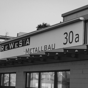 Detailansicht des Eingangsbereichs von Rewesta Metallbau mit Firmenschild, Hausnummer 30a und rotem Vordach mit Lochblechträgern.