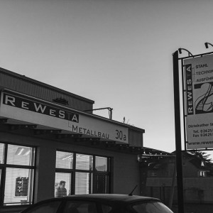 Außenansicht des Firmengebäudes von Rewesta Metallbau in Düren bei Sonnenuntergang, mit beleuchtetem Firmenschild und großem Werbeschild mit Kontaktinformationen.